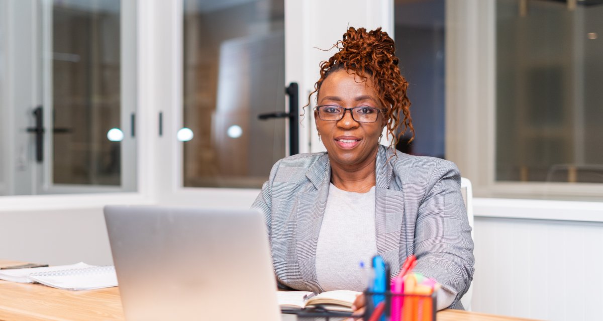Mature black woman sitting at desk wearing glasses looking at camera with laptop on the table.