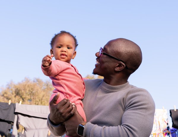 Black father holding daughter and smiling in the sun with clothe hanging in the background.