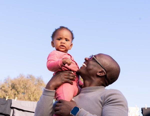 Black father smiling with daughter wearing pink outside with blue sky in the background