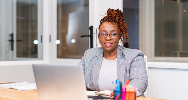 Mature black woman sitting at desk wearing glasses looking at camera with laptop on the table.