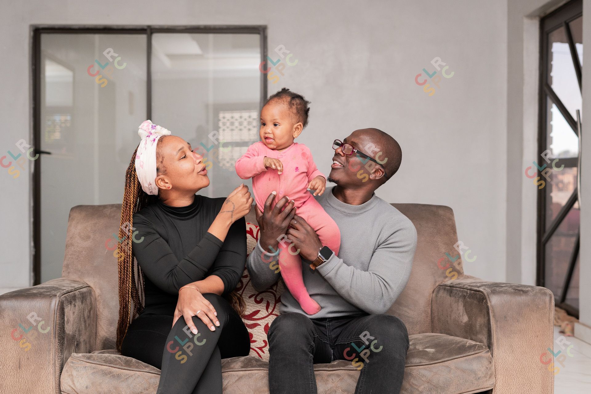 Black family smiles at home. Dad, Mom in black, and their 1-year-old daughter in pink.