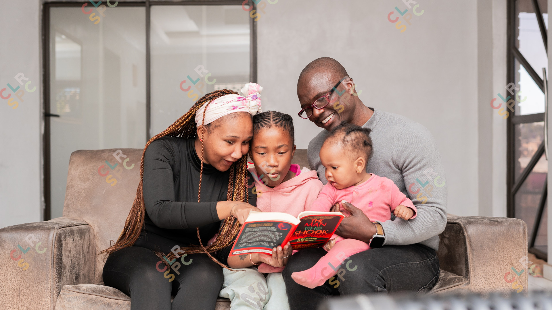 Loving black family reading a book at home on the couch, mom wearing black and kids wearing pink.