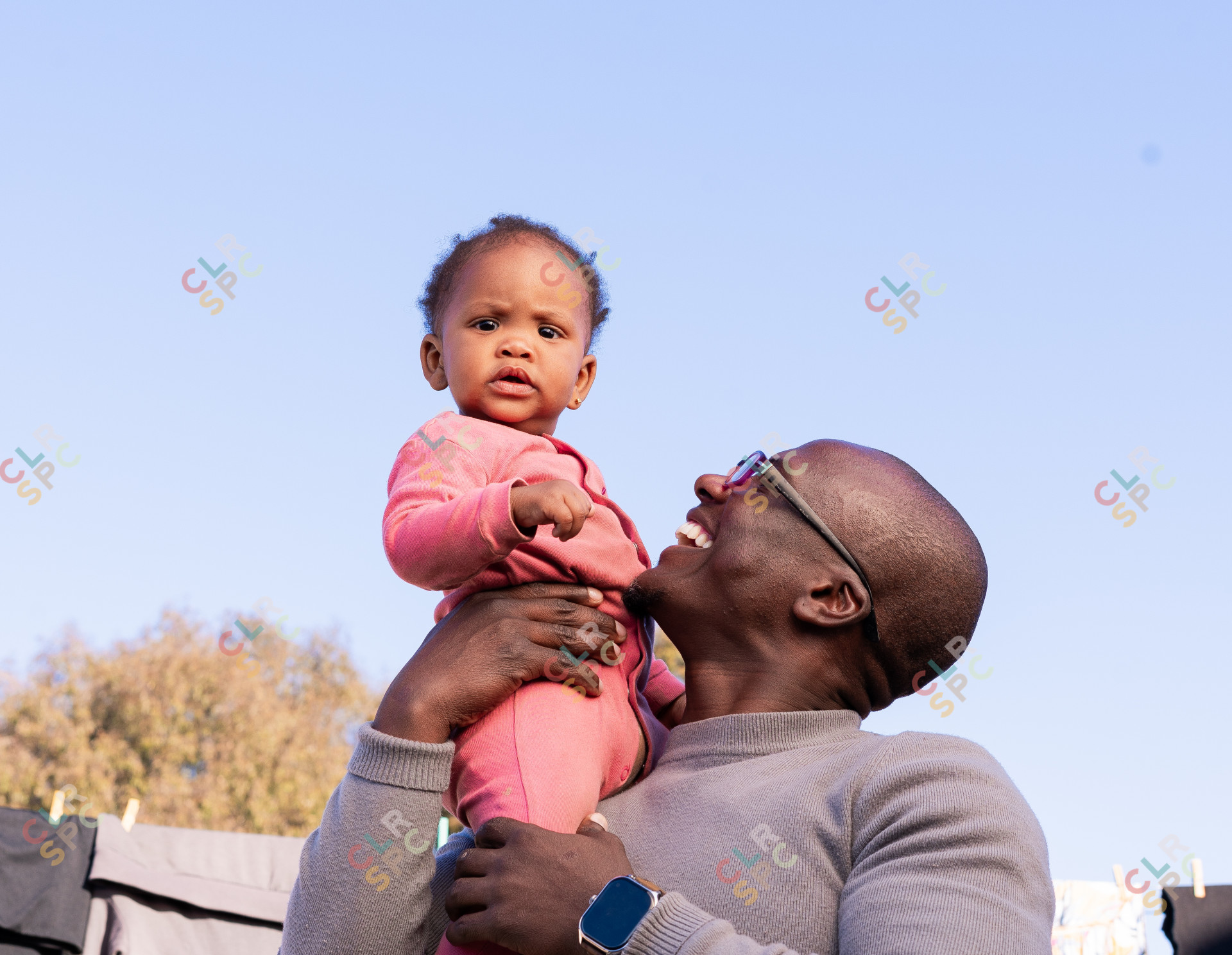 Black father smiling with daughter wearing pink outside with blue sky in the background
