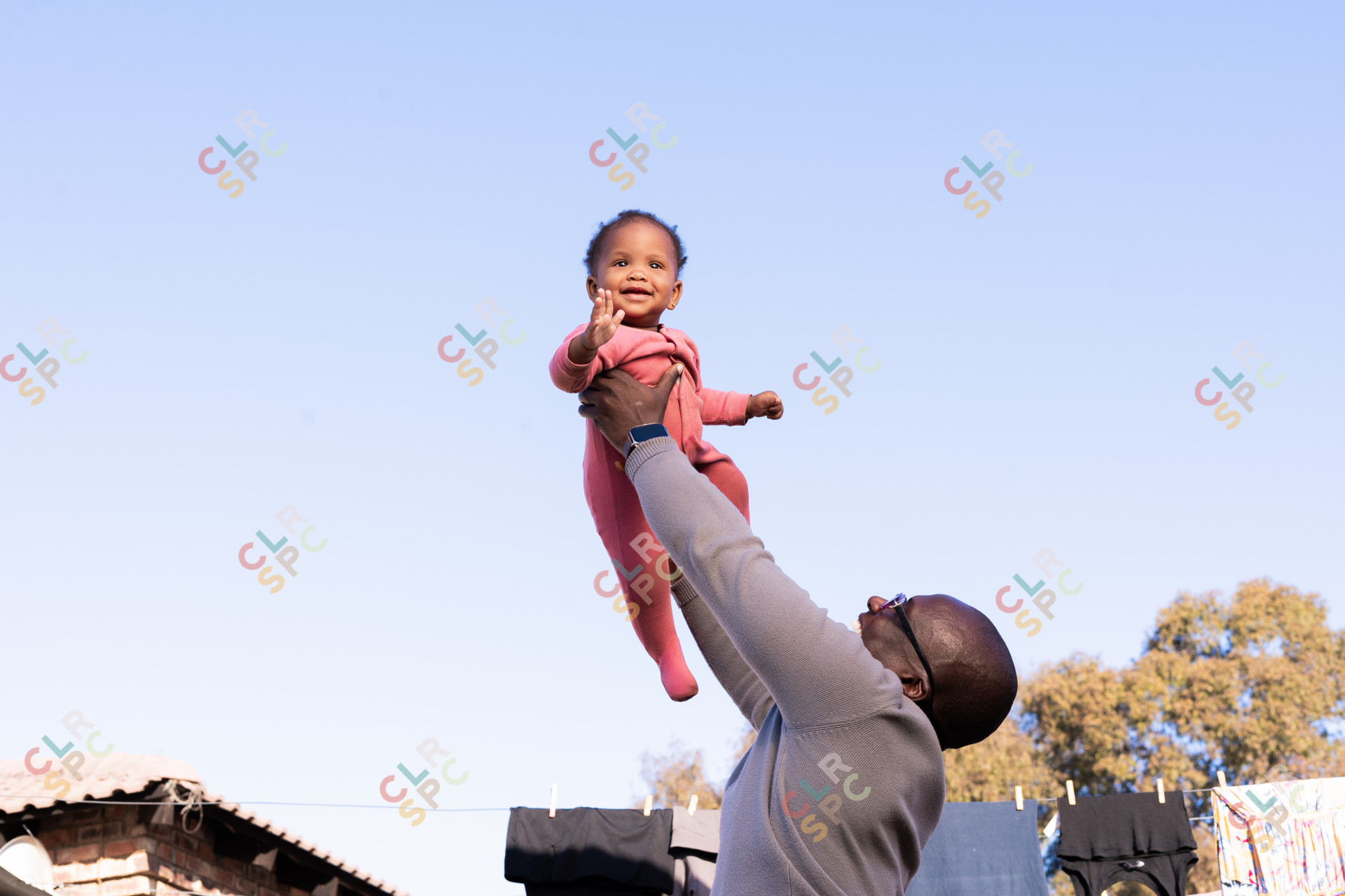 Black daughter smiling with her father outdoors wearing pink with clothes and trees in the background.