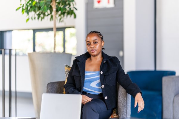 Young black woman sitting on the couch at work looking at the camera and a laptop in front of her.
